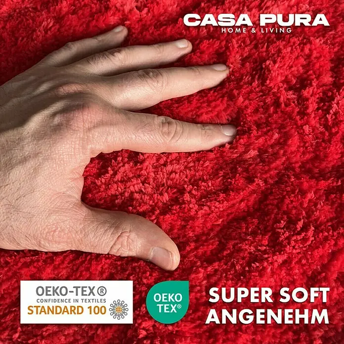 Floordirekt Badteppich Sky - 60 x 50 cm, Beerenrot, 100% Polyester - Nahaufnahme eines roten Casa Pura Teppichs mit Hand zur Hervorhebung seiner Weichheit, zertifiziert mit OEKO-TEX® Siegel.