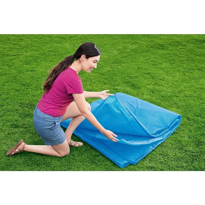Bestway Pool-Abdeckplane Solar - Durchmesser: 462 cm, Material: Kunststoff - Frau kniet auf Rasen und breitet eine blaue Poolabdeckung aus. Die Abdeckung ist aus Noppenfolie und glänzt.