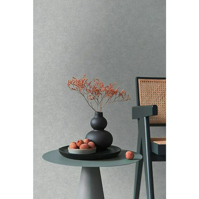 A.S. Création Metropolitan Stories Hot Spots Vliestapete - Grau-Blau, Uni, 10,05 x 0,53 m - Elegantes Stillleben mit einer schwarzen Vase, Trockenblumen und einer Schale mit Litschis auf einem runden Tisch neben einem stilvollen Stuhl.