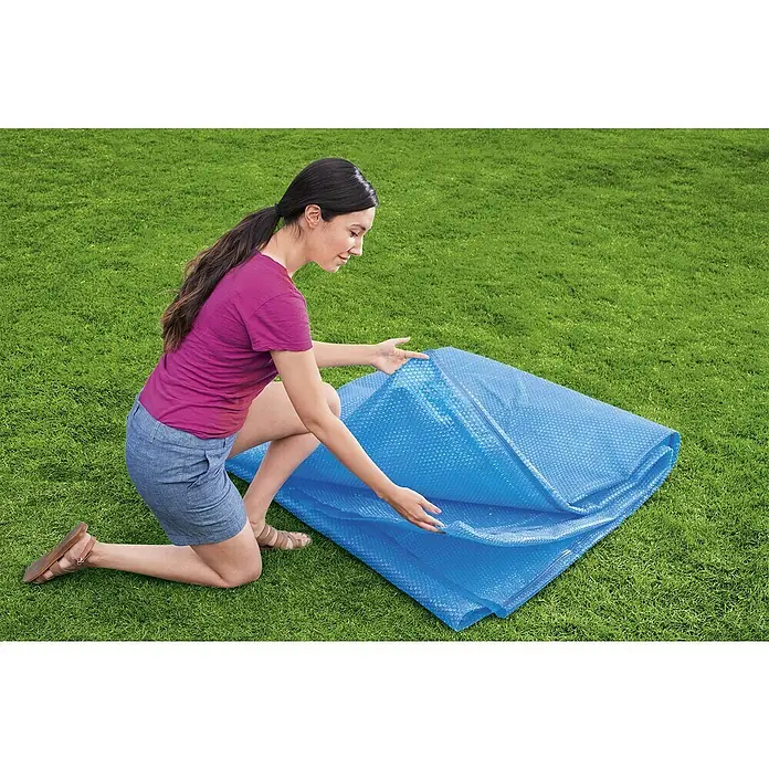 Bestway Solarabdeckplane - L x B: 336 x 703 cm, Material: Kunststoff - Frau kniet auf dem Rasen und entfaltet eine blaue Solarplane für den Pool, die mit Luftblasen versehen ist.