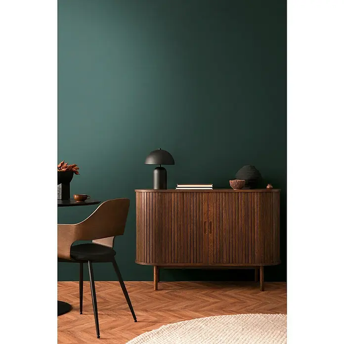 A.S. Création Pintwalls 2 Vliestapete - Grün, Uni, 10,05 x 0,53 m - Elegantes Sideboard aus Holz mit geriffelten Türen, verziert mit einer schwarzen Lampe, Büchern und dekorativen Schalen vor einer tiefgrünen Wand.