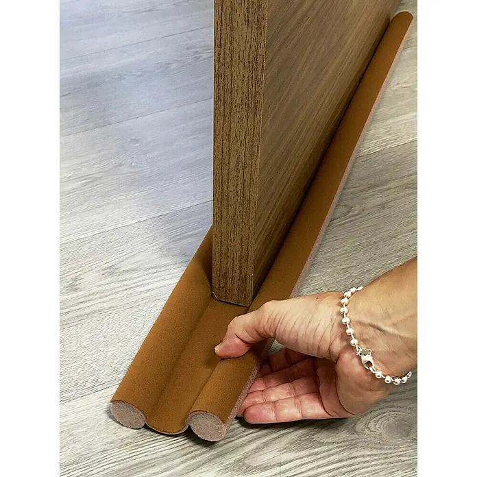 Junta para puerta Rodillo - Marrón, Largo: 95 cm - Primer plano de un burlete marrón debajo de una puerta de madera, ajustado por una mano, sobre un piso laminado gris.