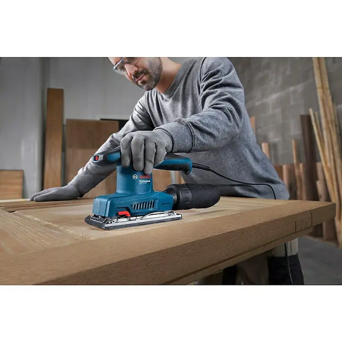 Bosch Professional Elektro-Schwingschleifer GSS 20-18 A -  - Ein Mann schleift Holz mit einem blauen Bosch Exzenterschleifer, wobei er Handschuhe und eine Schutzbrille trägt.
