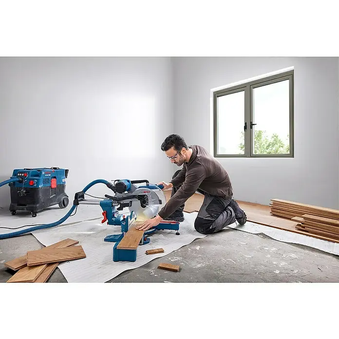Bosch Professional Kapp- & Gehrungssäge GCM 340-305D - 1.800 W, Durchmesser Sägeblatt: 305 mm, Leerlaufdrehzahl: 4.050 U/min - Ein Mann kniet vor einer Bosch Kapp- und Gehrungssäge, die an einen Staubsauger angeschlossen ist, um Holzböden zu schneiden.