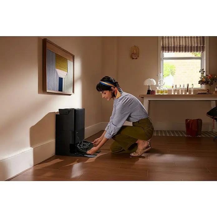 Roborock Saugwischroboter QR 598 Black - Saugkraft: 8.000 Pa, Akkulaufzeit: 180 min, Fassungsvermögen Roboter: 330 ml, Schwarz - Eine Frau interagiert mit einer schwarzen Roboterstaubsauger-Basisstation auf einem Holzboden in einem gemütlichen Zimmer.