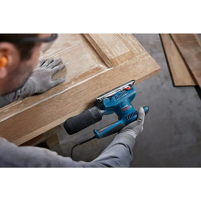Bosch Professional Elektro-Schwingschleifer GSS 20-18 A -  - Nahaufnahme eines Bosch-Exzenterschleifers beim Schleifen einer Holzoberfläche, getragen von einer Person mit Schutzhandschuhen.