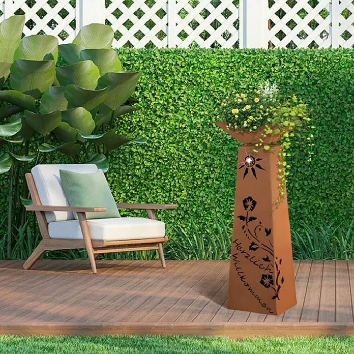 Jabo-Design Dekosäule mit Schale - 38 x 114 cm, Edelrost, Lilienranke - Das Bild zeigt einen dekorativen Pflanzturm mit Blumen in einem Garten, der eine warme und einladende Atmosphäre schafft.