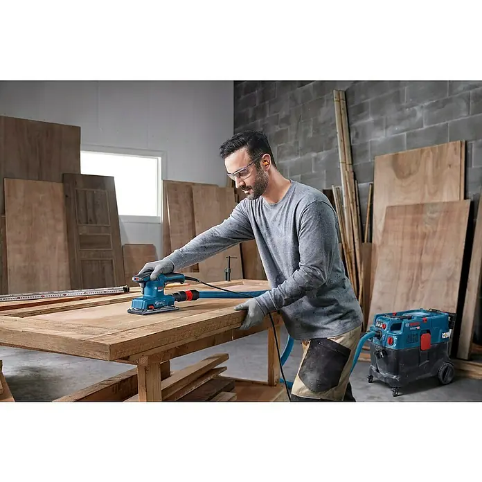 Bosch Professional Elektro-Schwingschleifer GSS 20-18 A -  - Ein Mann schleift mit einem Bosch-Schleifgerät ein Holzbrett in einer Werkstatt, umgeben von Holzmaterialien und einer Absauganlage.