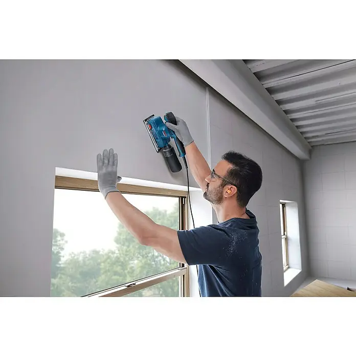Bosch Professional Elektro-Schwingschleifer GSS 20-18 A -  - Ein Mann benutzt einen blauen Bosch Schwingschleifer, um eine Wand über einem Fenster zu schleifen, wobei er graue Handschuhe trägt.