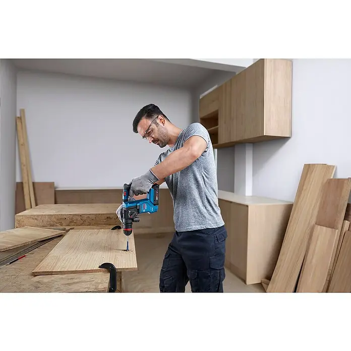 Bosch Professional AMPShare 18V Akku-Bohrhammer GBH 18V-18X - 18 V, Ohne Akku, 1,5 J - Ein Mann bohrt mit einer blauen Bosch-Bohrmaschine ein Loch in ein Holzbrett, während er Schutzbrille und Handschuhe trägt.