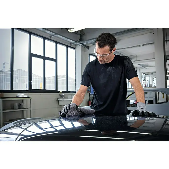 Bosch Professional Akku-Poliermaschine GPX 12V-125 - 12 V, Ohne Akku, Leerlaufdrehzahl: 1.400 U/min - 7.000 U/min - Mann mit Schutzbrille, Handschuhen und T-Shirt reinigt ein Auto mit einem Tuch. Die Spiegelung des Fensters ist auf dem Autolack zu sehen.