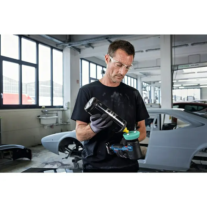 Bosch Professional Akku-Poliermaschine GPO 12V-77 - 12 V, Ohne Akku, Leerlaufdrehzahl: 600 U/min - 2.200 U/min - Ein Mann mit Schutzbrille trägt Politur auf eine Akku-Poliermaschine auf, im Hintergrund eine Autowerkstatt.