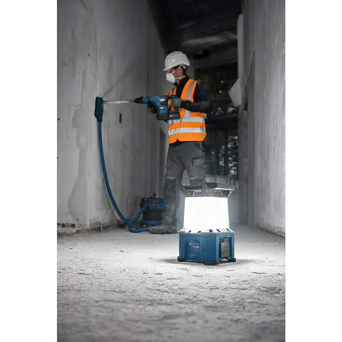 Bosch Professional AMPShare 18V Akku-Bauleuchte - 18 V, Anzahl Akkus: Ohne Akku, 7.500 lm - Ein Bauarbeiter mit Schutzhelm und Staubmaske bohrt mit einem Bosch Bohrhammer ein Loch in eine Wand, beleuchtet von einem Bosch Baustrahler.