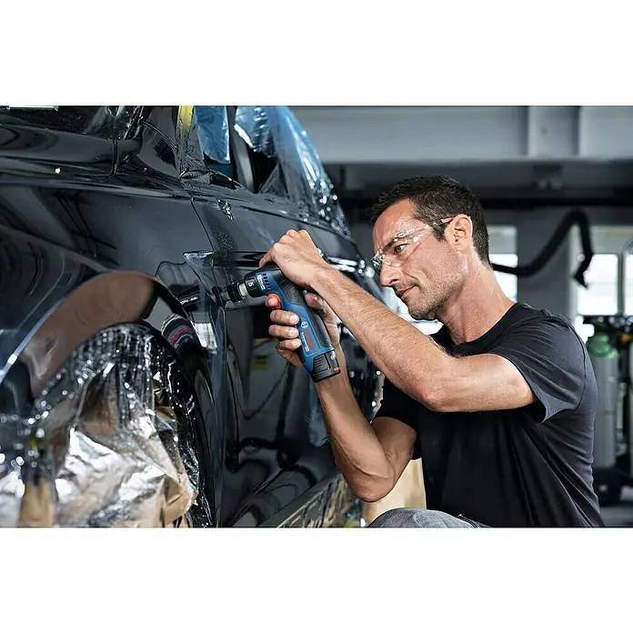 Bosch Professional Akku-Exzenterschleifer GEX 12V-32 - 12 V, Ohne Akku, Durchmesser Schleifteller: 32 mm - Ein Mann verwendet einen Bosch Professional Akku-Bohrschrauber an einem schwarzen Auto, um eine Schutzfolie anzubringen. Er trägt dabei eine Schutzbrille.
