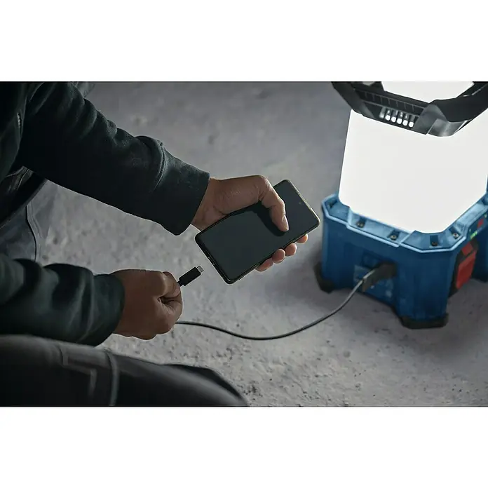 Bosch Professional AMPShare 18V Akku-Bauleuchte - 18 V, Anzahl Akkus: Ohne Akku, 7.500 lm - Das Bild zeigt eine Bosch Akku-Baustellenleuchte, die ein Smartphone auflädt. Die Lampe ist blau und hat ein helles, weißes Licht.