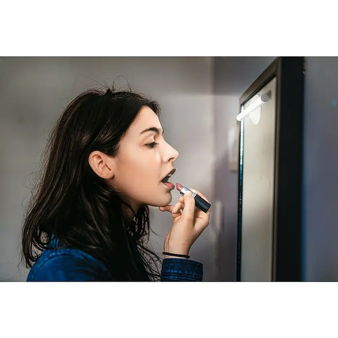 Osram LED-Unterbauleuchte Schrankleuchte LEDStixx - L x B x H: 20,6 x 1,7 x 1,7 cm, Lichtfarbe: Kaltweiß - Eine Frau trägt Lippenstift auf, während sie in den Spiegel schaut. Der Fokus liegt auf dem Lippenstift und der Schönheitspflege.