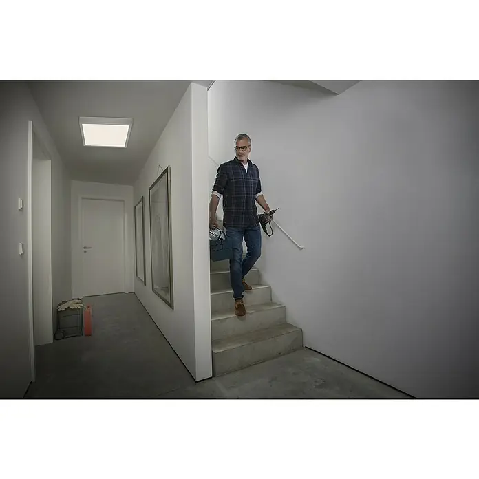 Ledvance LED-Panel Frame Essential - Weiß, Passend für: Osram Essential LED-Panel 60 x 60 cm - Ein Mann steigt mit Werkzeug die Treppe in einem hellen Flur hinunter, Deckenleuchte erhellt den Raum.\n