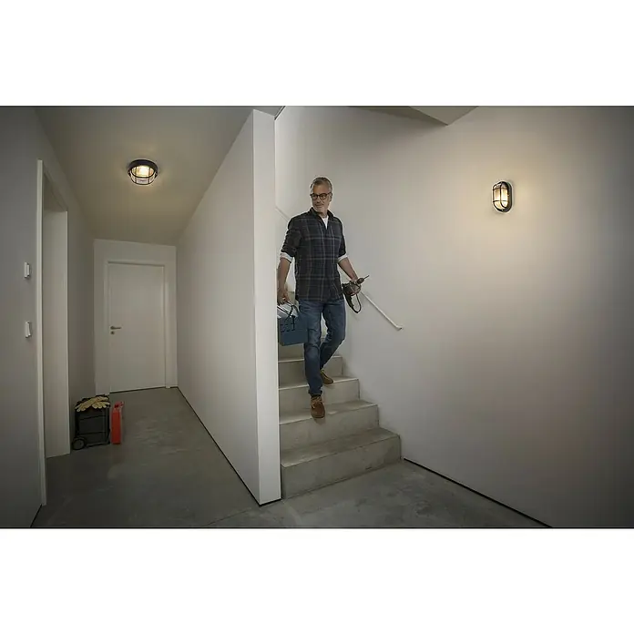 Osram Wand- & Deckenleuchte Bulkhead Essential - L x B x H: 9,8 x 11 x 20,7 cm, Schwarz, E27 - Ein Mann steigt eine Treppe in einem Flur hinunter, der mit modernen Wand- und Deckenleuchten beleuchtet wird, die ein warmes, einladendes Licht spenden.