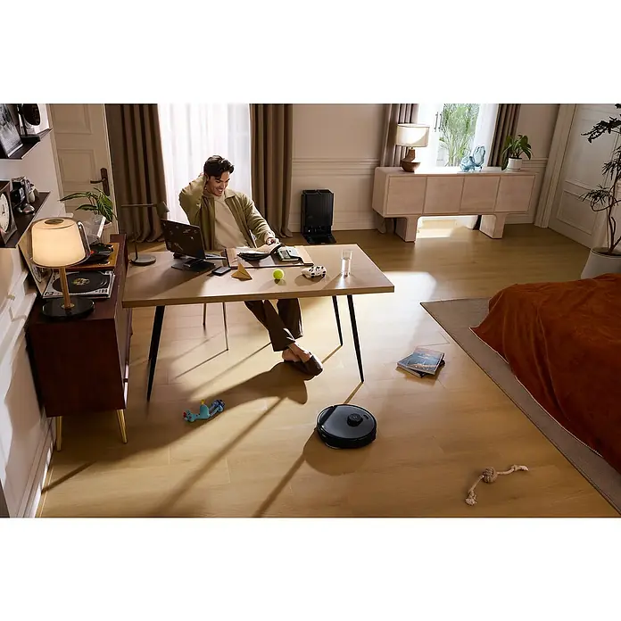 Roborock Saugwischroboter Qrevo Edge 5V1 Black - Saugkraft: 18.500 Pa, Akkulaufzeit: 180 min, Fassungsvermögen Roboter: 325 ml, Schwarz - Bild von einem Mann, der an einem Tisch sitzt, während ein Roboterstaubsauger den Boden reinigt. Fokus liegt auf dem Roboterstaubsauger in Aktion.