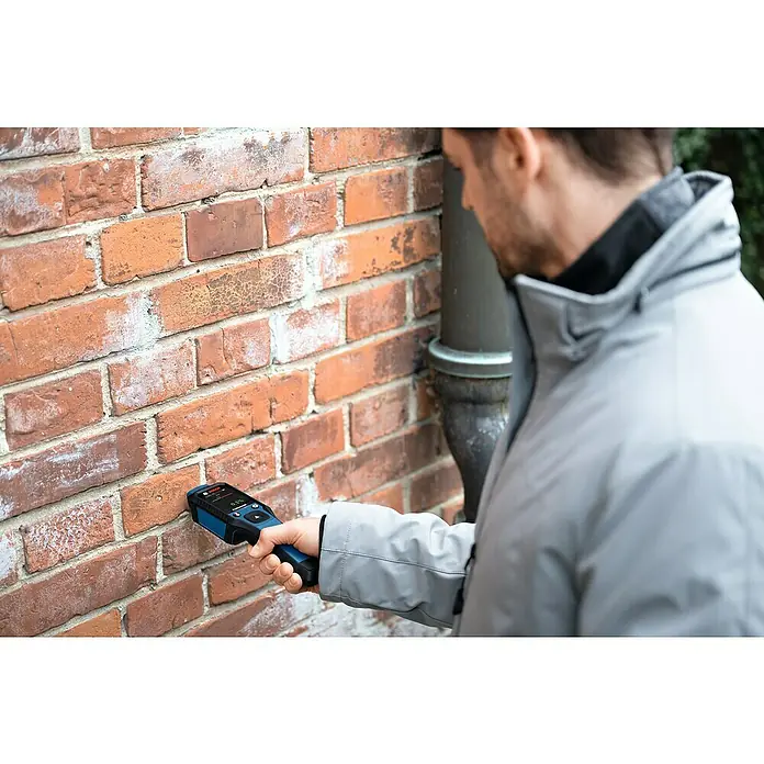 Bosch Feuchtigkeitsmessgerät GMP 2-15 - Messgenauigkeit: Umgebungstemperatur typisch: +/- 1 °C, Mit Batterien - Ein Mann benutzt ein Bosch-Messgerät an einer Ziegelwand, um Feuchtigkeit zu messen. Das Gerät ist blau und schwarz.