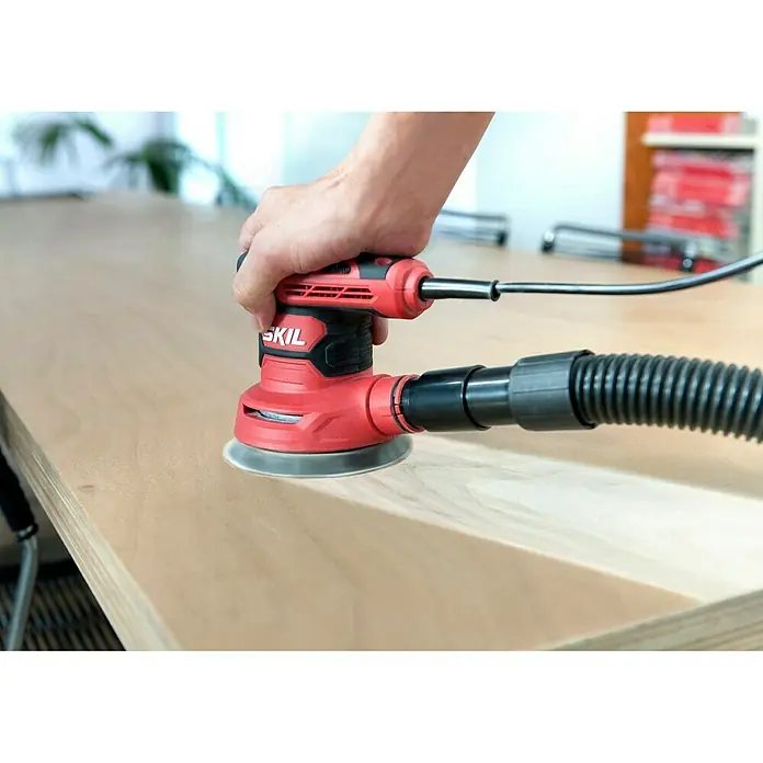 Skil Elektro-Exzenterschleifer 7471 AA - 300 W, Durchmesser Schleifteller: 125 mm - Nahaufnahme eines roten SKIL-Exzenterschleifers mit Staubabsaugung beim Schleifen einer Holzoberfläche.