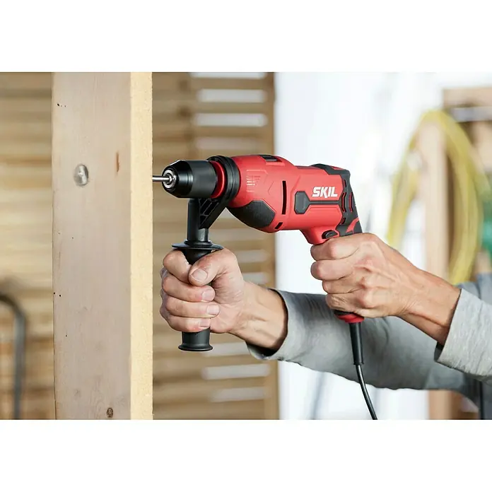 Skil Schlagbohrmaschine 6725 AA - 710 W, Schnellspannbohrfutter - Nahaufnahme eines roten Skil Bohrhammers, der von einer Person gehalten wird, während er in ein Holzstück bohrt.