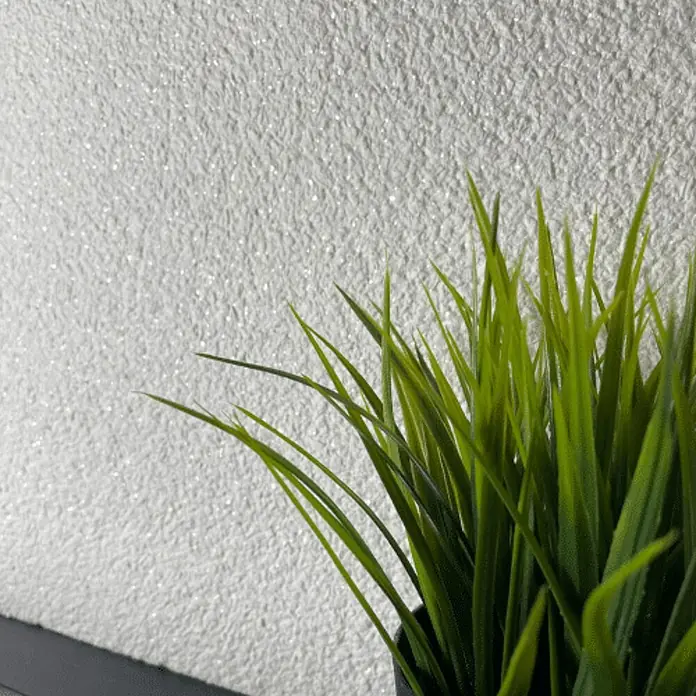 Floxxan Baumwollputz Montana 512 Cellulose - Putz Tapete Flüssigtapete Weiss m. Mica fein - Weiss mit Mica fein, 1 kg, Lösemittelfrei - Nahaufnahme von künstlichem Gras vor einem schimmernden Hintergrund. Die grünen Halme bilden einen lebendigen Kontrast zur glitzernden Oberfläche.
