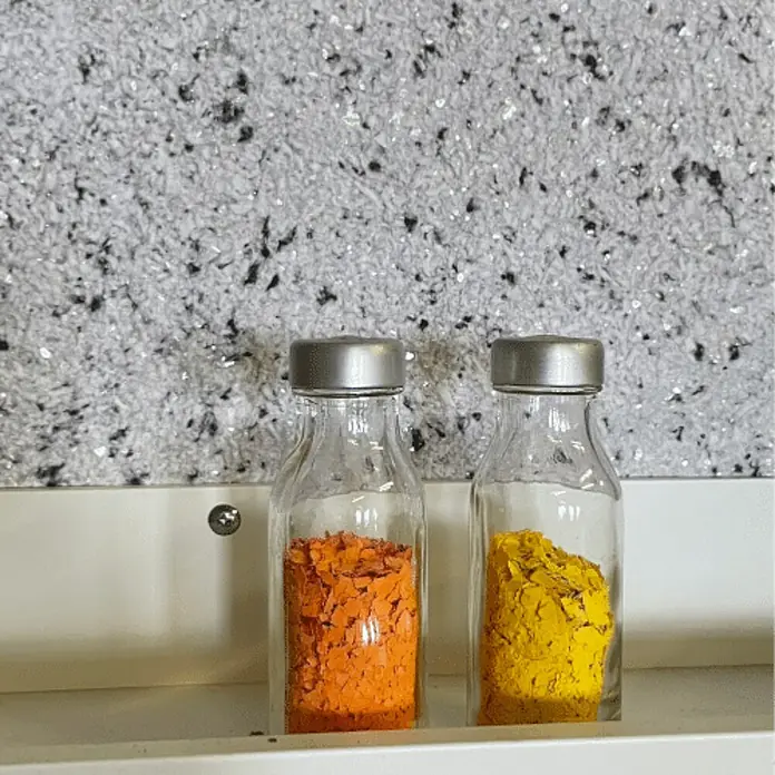 Floxxan Baumwollputz California 201 - Putz Tapete Flüssigtapete Anthrazit m. Mica Natur - Anthrazit, 1 kg, Lösemittelfrei - Nahaufnahme von zwei kleinen Glasflaschen mit Schraubverschluss, die mit bunten Flocken gefüllt sind, darunter Orange und Gelb.