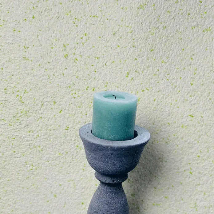 Floxxan Baumwollputz Montana 515 Cellulose - Putz Tapete Flüssigtapete Lemon Green - Lemon Green, 1 kg, Lösemittelfrei - Kerzenhalter mit einer blauen Kerze darauf, vor einer hellen Wand. Das minimalistische Design verleiht eine moderne Note.
