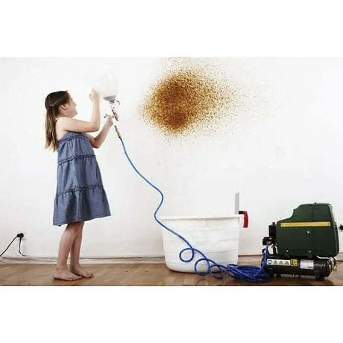Floxxan Baumwollputz Trichterspritzpistole für die Verarbeitung von Baumwollputz / Flüssigtapete -  - Ein Mädchen in einem blauen Kleid, das mit einer Spritzpistole Farbe an eine Wand sprüht, daneben ein Farbbehälter und ein Kompressor.