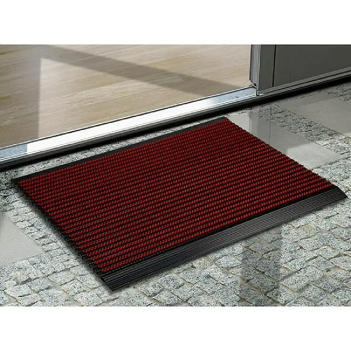 Fußmatte Power Brush - Rot, 60 x 40 cm, 100 % Nylon - Eine rote Fußmatte mit schwarzem Rand liegt vor einer Tür auf einem gepflasterten Untergrund.