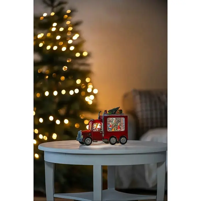 Konstsmide LED-Weihnachtsdekofigur LKW - 28 x 9,7 x 21,5 cm, Rot, Batteriebetrieben, LED - Weihnachtliche Szene mit einem dekorativen, roten Spielzeug-Truck mit Schneekugel auf einem Tisch, vor einem Weihnachtsbaum mit Lichtern.