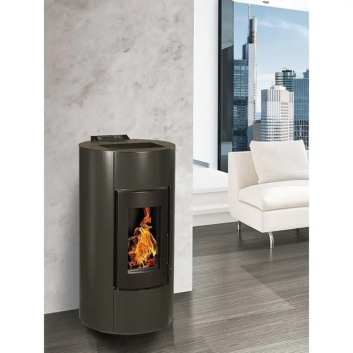 Pelletofen Angelina - 11,4 kW, Raumheizvermögen: 320 m³, 0,73 kg/h - 2,5 kg/h, Farbe Verkleidung: Schwarz - Ein moderner Pelletofen mit Flammen steht vor einer grauen Wand. Im Hintergrund ist ein Sofa und eine Fensterfront mit Blick auf Hochhäuser zu sehen.