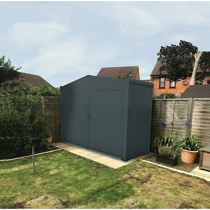 Wismar-NFI Gerätehaus Aster 2V - Außenmaß inkl. Dachüberstand (B x T): 325 x 125 cm, Stahl, Anthrazit/Weiß, Doppeltür - Grauer Metallschuppen mit Doppeltüren im Garten, umgeben von Rasen, Pflanzen und einem Holzzaun.