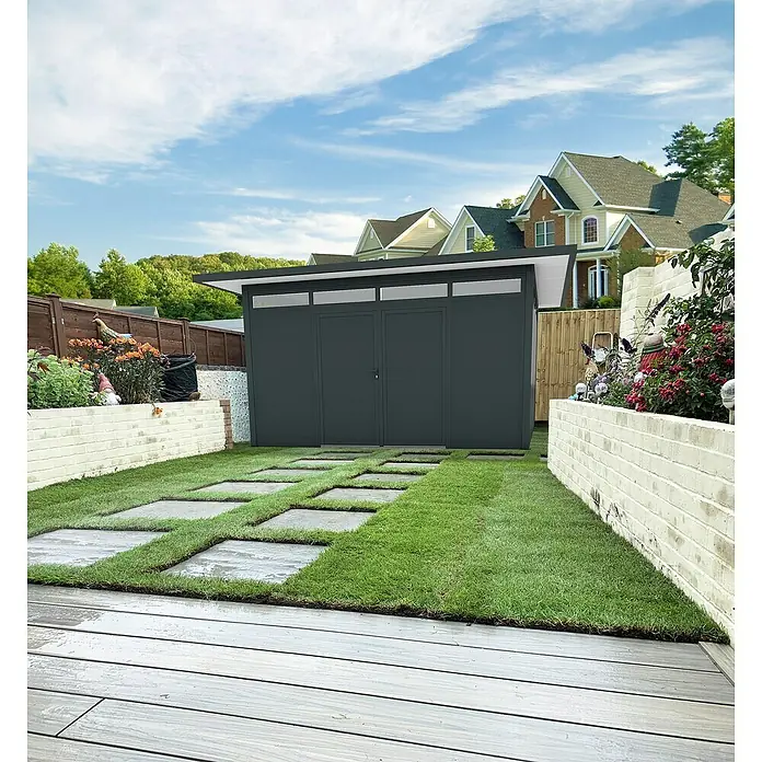 Wismar-NFI Gerätehaus Trefle 44 - Außenmaß inkl. Dachüberstand (B x T): 525 x 525 cm, Stahl, Anthrazit/Weiß - Moderne Gartenlaube mit dunklen Wänden und begrünten Trittsteinen vor einem begrünten Garten mit Häusern im Hintergrund.