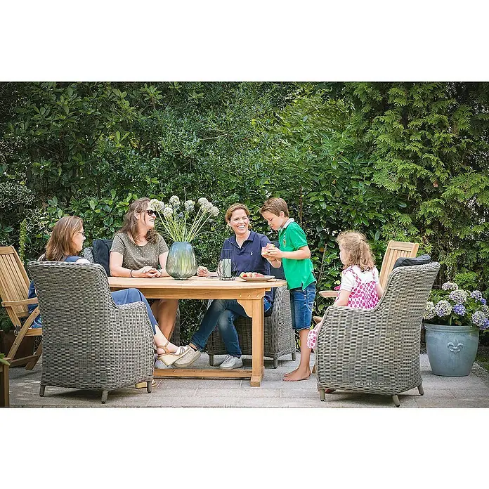 Kai Wiechmann Gartentisch - L x B x H: 240 x 120 x 75 cm, Teakholz, Oval, Naturbelassen - Gartenmöbel-Szene mit Holztisch und Korbstühlen, umgeben von üppigem Grün, ideal für entspannte Stunden im Freien.