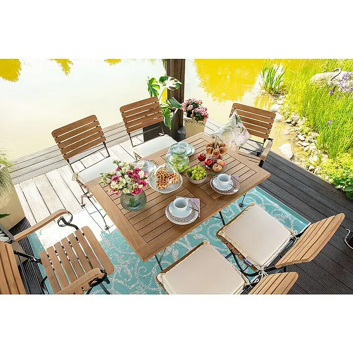 Kai Wiechmann Gartentisch - L x B x H: 150 x 80 x 72 cm, Teakholz, Rechteckig, Naturbelassen - Ein quadratischer Gartentisch aus Holz mit Stühlen steht auf einem blauen Teppich, dekoriert mit Blumen, Geschirr und Speisen, für eine entspannte Mahlzeit im Freien.