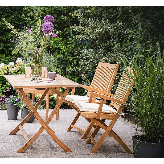 Kai Wiechmann Gartentisch - L x B x H: 70 x 70 x 75 cm, Teakholz, Quadratisch, Naturbelassen - Gartenszene mit einem quadratischen Holztisch und zwei passenden Stühlen mit weißen Sitzkissen vor üppigem Grün.