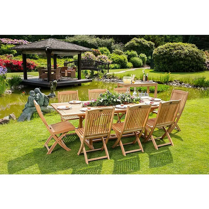 Kai Wiechmann Gartentisch - L x B x H: 120 x 45 x 90 cm, Teak, Rechteckig, Naturbelassen - Ein Gartentisch aus Holz mit Stühlen, dekoriert mit Blumen, steht auf einer grünen Wiese in der Nähe eines Teichs.