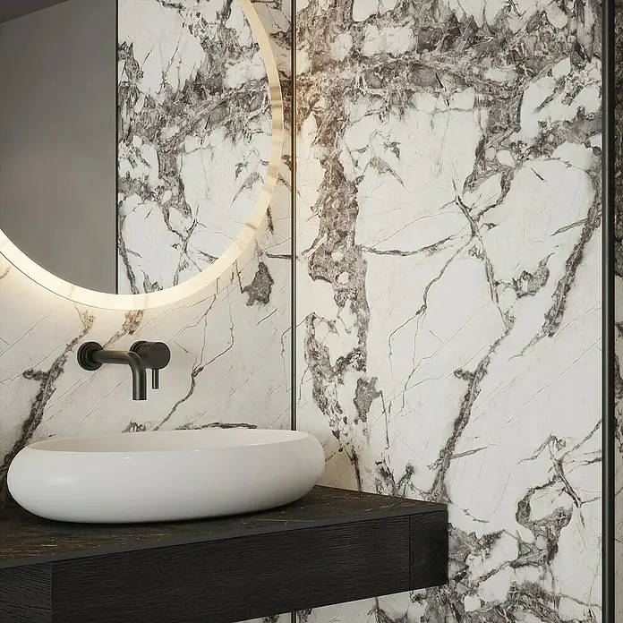 Wetwall Wandpaneel Alloy - 90 x 244 cm, Calacatta Fantasia - Elegantes Badezimmer-Design mit einem runden Spiegel mit Beleuchtung, einem stilvollen Aufsatzwaschbecken und dunkler Waschtischplatte vor einer Marmorwand.