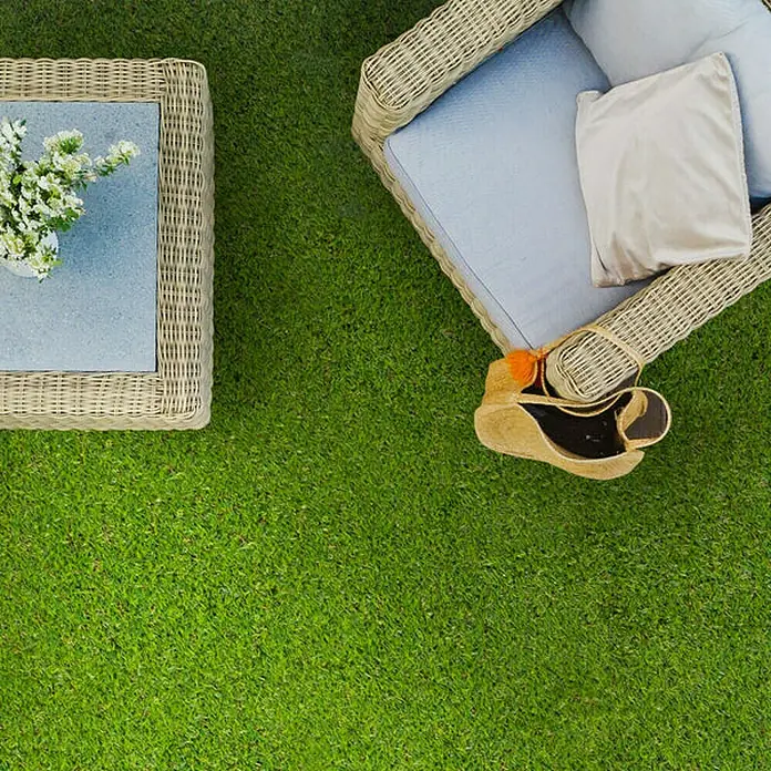 Floordirekt Klickfliese Kunstgras 30 x 30 cm - 30 x 30 x 3,5 cm, 1 Stk. - Nahaufnahme eines Gartensets mit einem Tisch mit Blumen und einem Korbsessel mit Kissen und einer Tasche auf grünem Rasen.