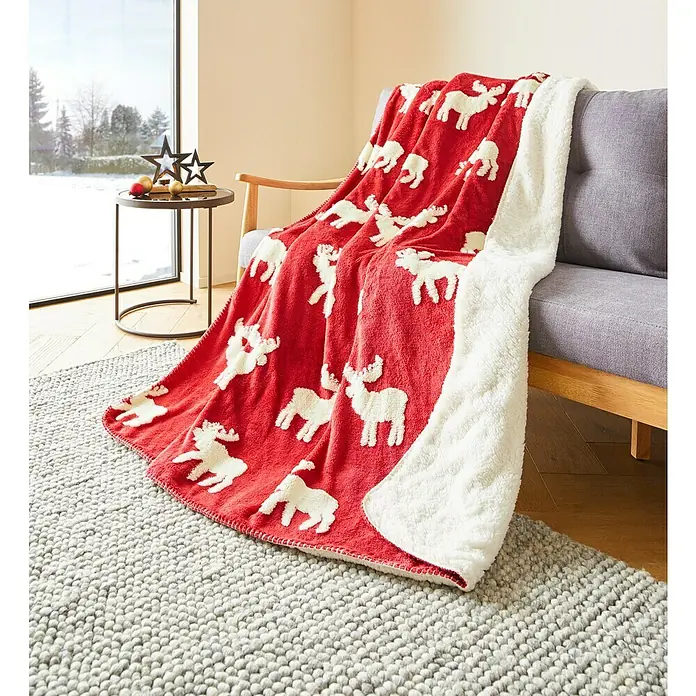 Decke Reindeer Xmas - Rot/Weiß, 180 x 140 cm, 100 % Polyester - Rote Kuscheldecke mit weißen Rentieren auf einem grauen Sofa, perfekt für gemütliche Winterabende. Die Rückseite ist aus flauschigem, weißem Sherpa-Fleece.