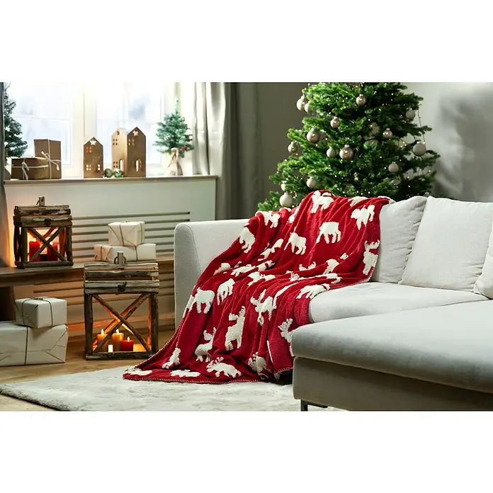 Decke Reindeer Xmas - Rot/Weiß, 180 x 140 cm, 100 % Polyester - Gemütliche Weihnachtsszene mit einer roten Decke mit weißen Elchen auf einem Sofa, neben einem Weihnachtsbaum und Geschenken.