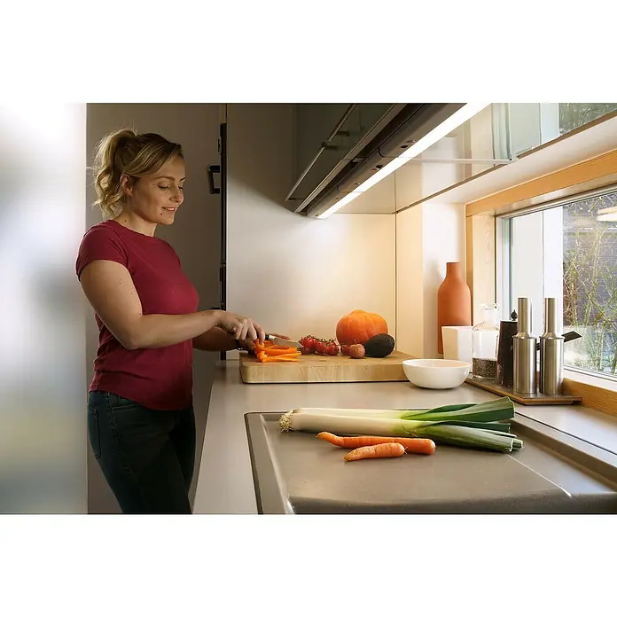 Osram Linear LED-Unterbauleuchte LED Flat - 6 W, Länge: 370 mm, Warmweiß - Frau bereitet in einer Küche mit Unterbauleuchten Gemüse zu. Die Küchenleuchten erhellen Arbeitsfläche und Lebensmittel.