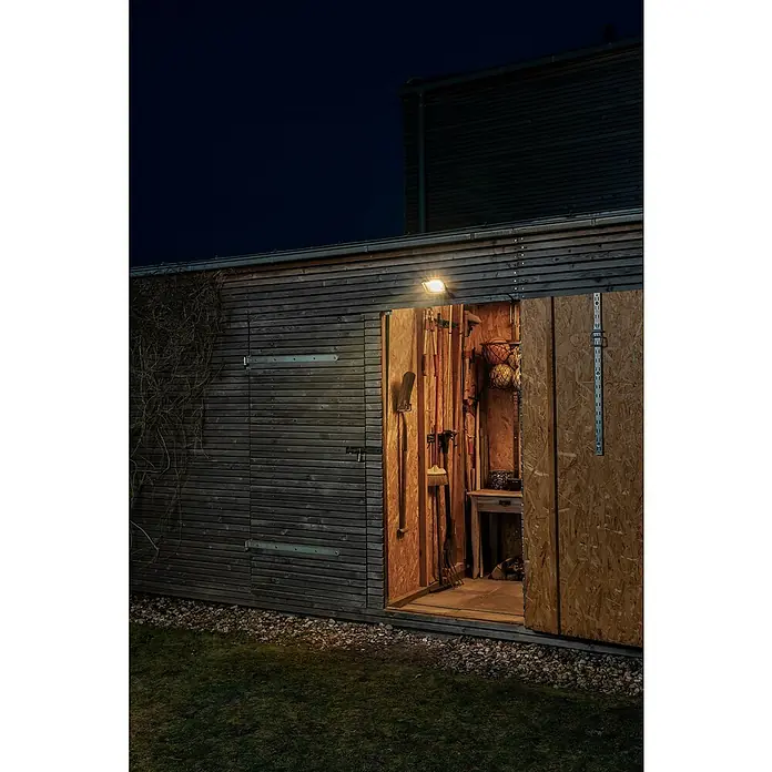 Osram LED-Strahler Endura Flood - 30 W, Anthrazit, Warmweiß, IP65 - Außenaufnahme eines Holzschuppens mit Gartengeräten und offenstehender Tür, der von einer Außenleuchte beleuchtet wird.