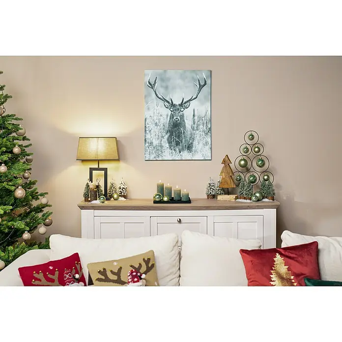 Stumpenkerzen-Set Rustic Safe - 4 -tlg., Smaragd - Festliche Wohnzimmerdekoration mit Weihnachtsbaum, Geweihbild, Lampe und dekorativen Elementen auf einem Sideboard.