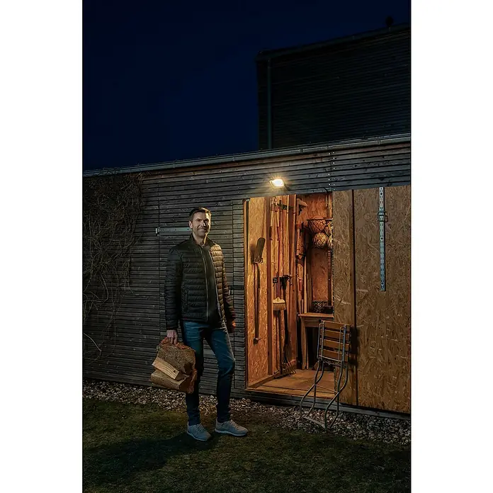 Osram LED-Strahler Endura Flood - 30 W, Anthrazit, Warmweiß, IP65 - Ein Mann steht mit Holz vor einem Schuppen, der mit einer Außenleuchte beleuchtet wird.