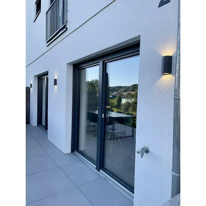 Ledvance Aplique exterior LED - 12 W, 5,5 cm x 9 mm x 16 cm, Gris oscuro, Claro, IP44 - Vista exterior de una casa moderna con puertas correderas grises e iluminación exterior, proyectando una luz cálida en la fachada.