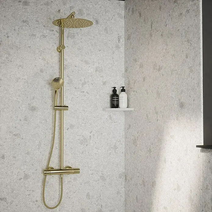 Wetwall Wandpaneel Elite - 16 x 242 cm, Terrazzo White, Abgerundete Kante - Nahaufnahme einer goldenen Duschgarnitur mit Kopf- und Handbrause vor einer grauen Wand mit zwei Pflegeprodukten auf einem Regal.