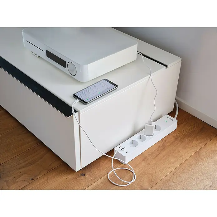 Osram Smart+ Smart-Steckdosenleiste 2. Gen. 2 USB-A, 2 USB-C - Weiß - Eine weiße OSRAM-Mehrfachsteckdose mit USB-Anschlüssen und einem Ladegerät, auf einem Holzboden liegend. Smartphone liegt oben.